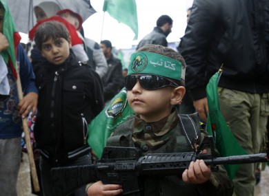 A Palestinian boy wears a green headband with an Arabic slogan and green Islamic flags while holding a toy gun.